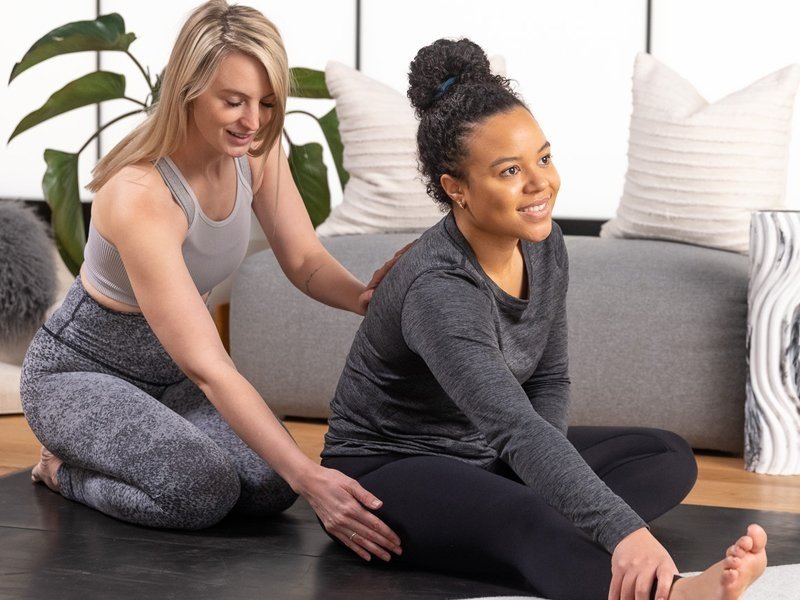 Trainer guiding woman through seated hamstring stretch on yoga mat.