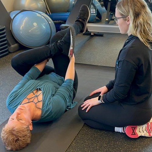 Trainer assisting client with lying hamstring stretch on a gym mat.