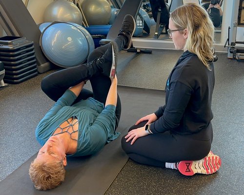 Trainer guiding client through lying hamstring stretch on gym mat.