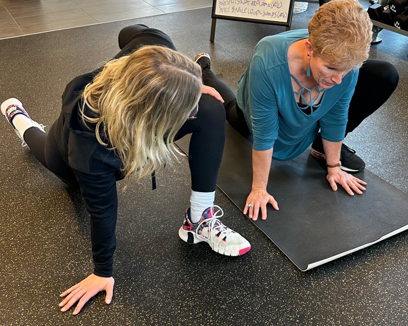 Trainer and client performing deep hip stretch together on gym floor.