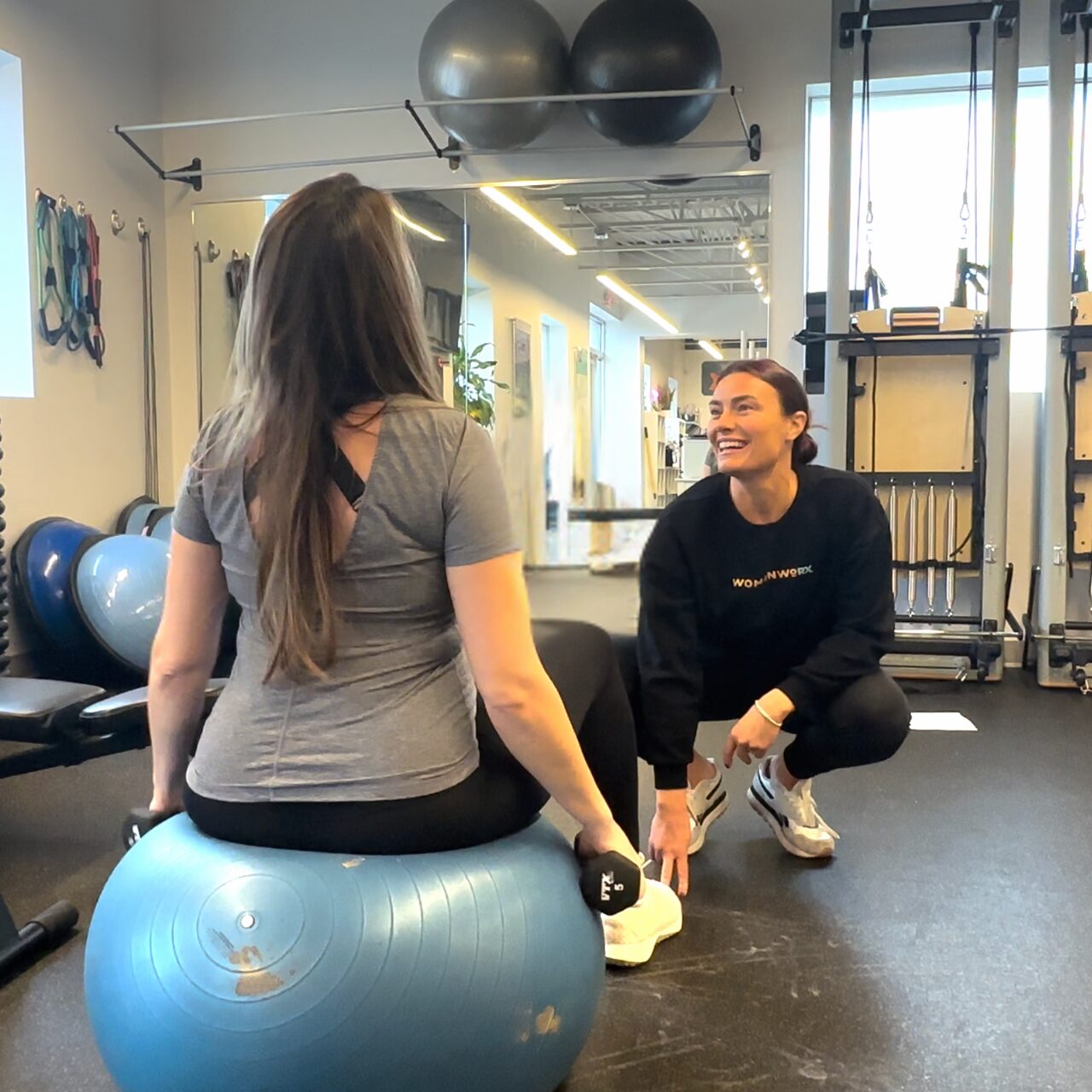 Trainer smiling while assisting woman seated on exercise ball holding dumbbells.
