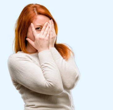 Woman with red hair playfully covering face with hands, smiling shyly.