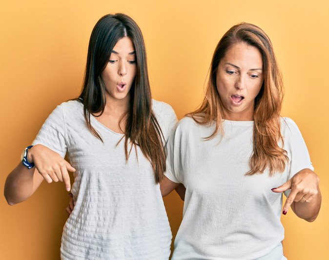 Two surprised women in white shirts pointing downward with shocked expressions.