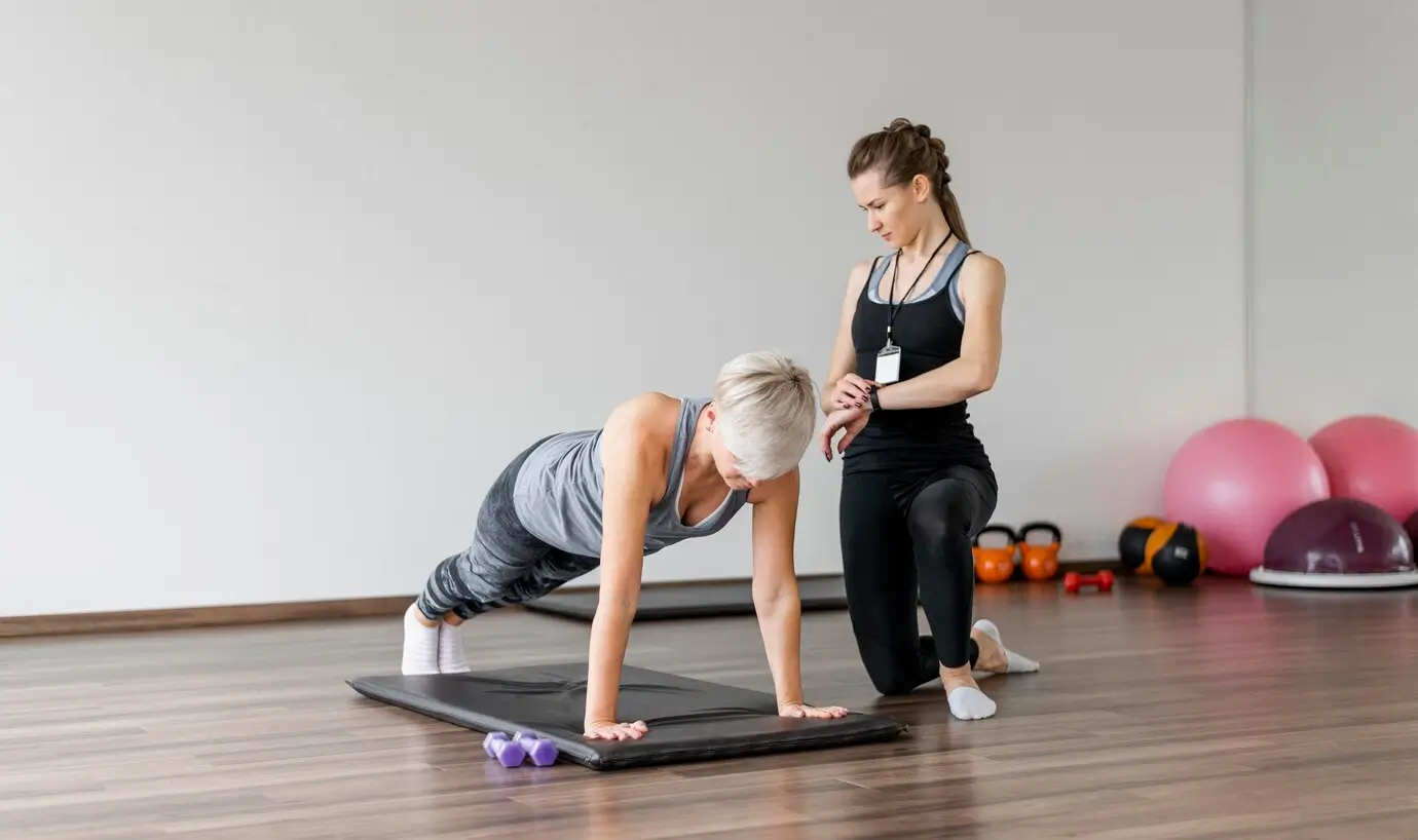 Older woman performing a plank exercise with guidance from a fitness trainer.